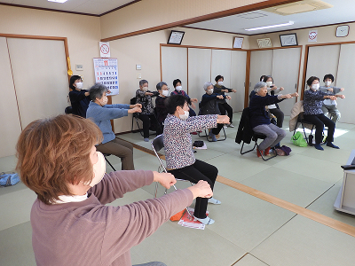13人の参加者が椅子に座り、両手を握りながら両腕を前に伸ばして体操しています。