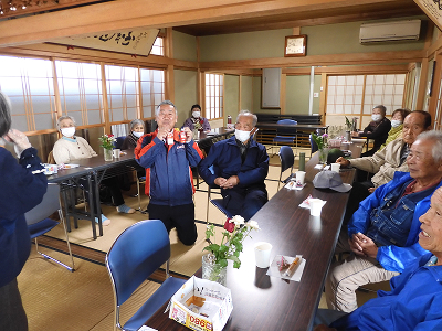 10人ほどの高齢の方がテーブルに座っています。うち一人が、膝立ちで明治R-1を両手に持って見せています。