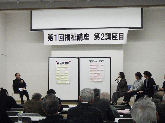左側のボードに「福祉事業所」と書かれた紙が貼られ、その中にたくさんの付箋が貼られています。右側のボードには「学生ボランティア」と書かれた紙が貼られ、その中にたくさんの付箋が貼られています。それらのボードの左側に男性一人が座り、右側に男女4人が座っています。左側の男性と右側の女性がマイクを持って話しています。