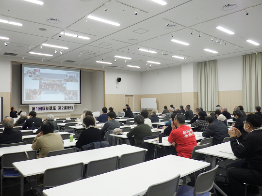 前方のプロジェクターを使い、男性が話をしています。大勢の人が座って話を聞いています。プロジェクターの下には「第１回福祉講座 第２講座目」と書かれた紙が貼られています。