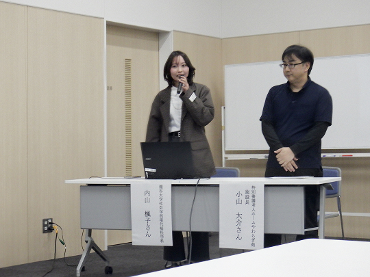 女性と男性がテーブルから立ち上がり、女性がマイクを持って話しています。女性の前には「内山楓子さん」、男性の前には「小山大介さん」とかかれた紙が貼られています。テーブルの上にはノートパソコンが置かれています。