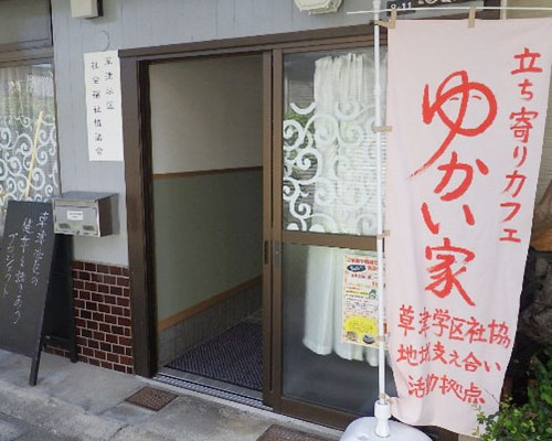 ゆかい家の玄関です。引き戸が開いています。傍にピンク色ののぼり旗が立てられており、赤い文字で「立ち寄りカフェゆかい家 草津学区社協地域支え合い活動拠点」と書かれています。
