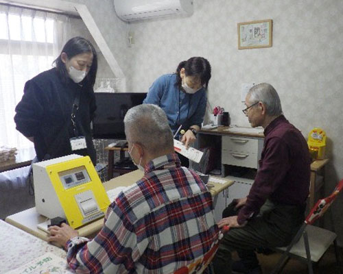 高齢男性２人がテーブルに座り、２人の女性が正面に立って話しています。テーブルには黄色い機械が置かれています。