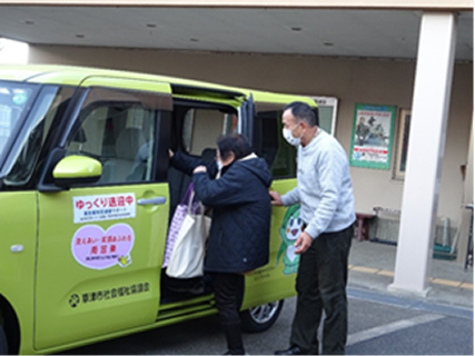 ご年配の女性が黄緑色の軽自動車に乗り込んでいます。送迎ボランティアの男性が傍で支えています。
