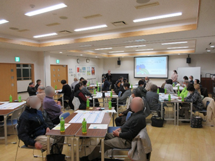 ご年配の方々が４、５人ずつに分かれてテーブルに座っており、前方のプロジェクターで説明している講師の話を聞いています。