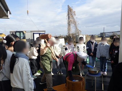 屋外でご年配の男女が餅つきを行っていて、その様子をたくさんの子供たちが取り囲んで注目しています。