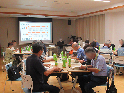 参加者が5人ずつ4テーブルに分かれて意見交換を行っています。前にはプロジェクターを使い説明する講師が3人座っています。